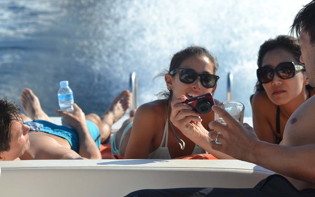 Guy showing two girls photos on a red digital camera while lounging on aft sundeck aboard a charter boat in Ibiza