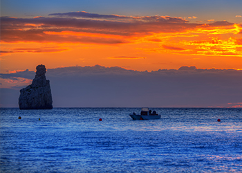 Boat passing the famous Benirras rocky outcrop at sunset in the sea off Benirras beach in northern Ibiza